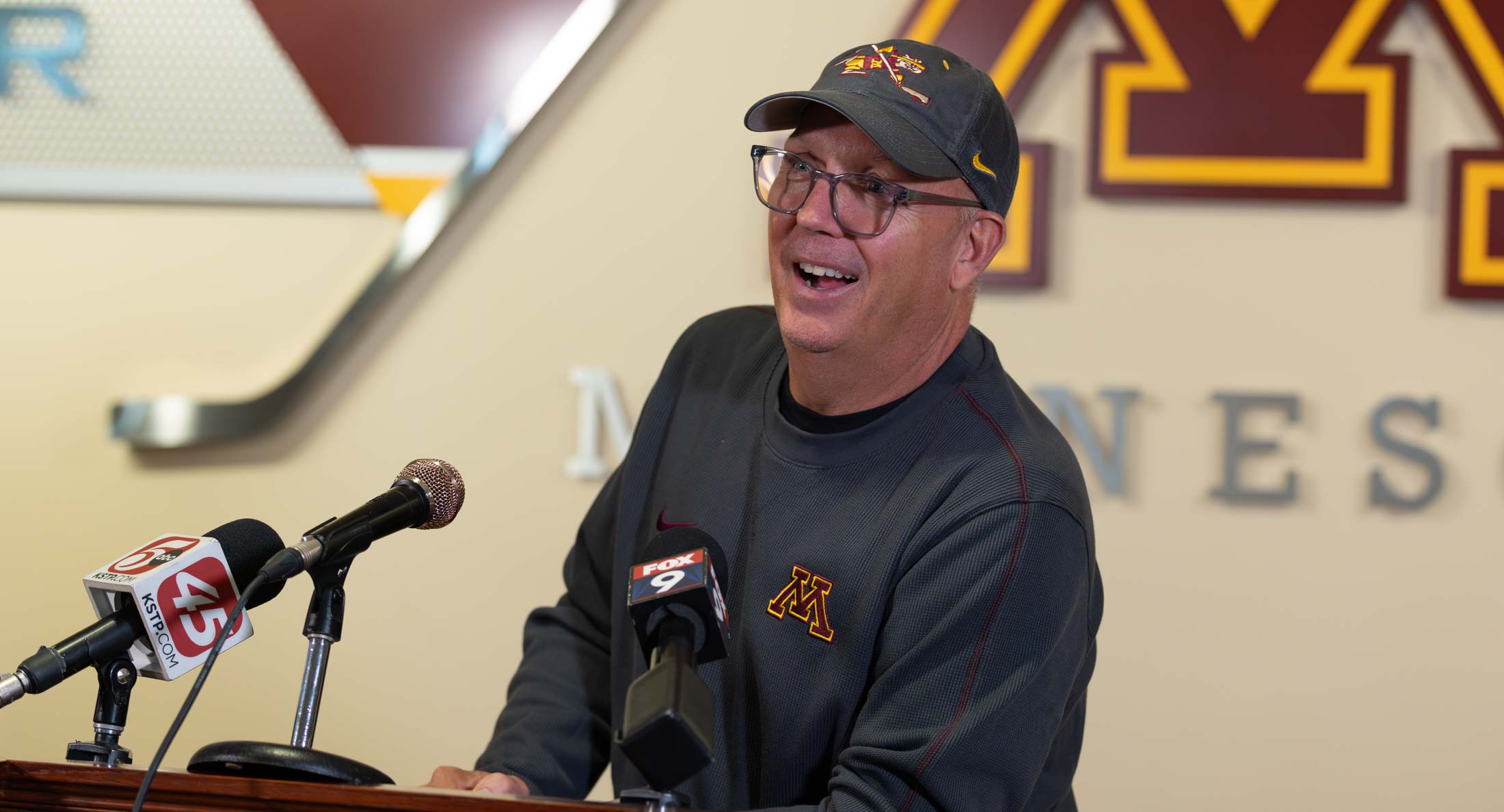 Bob Motzko wearing a Gophers hat and sweatshirt, smiling while speaking at a podium with microphones