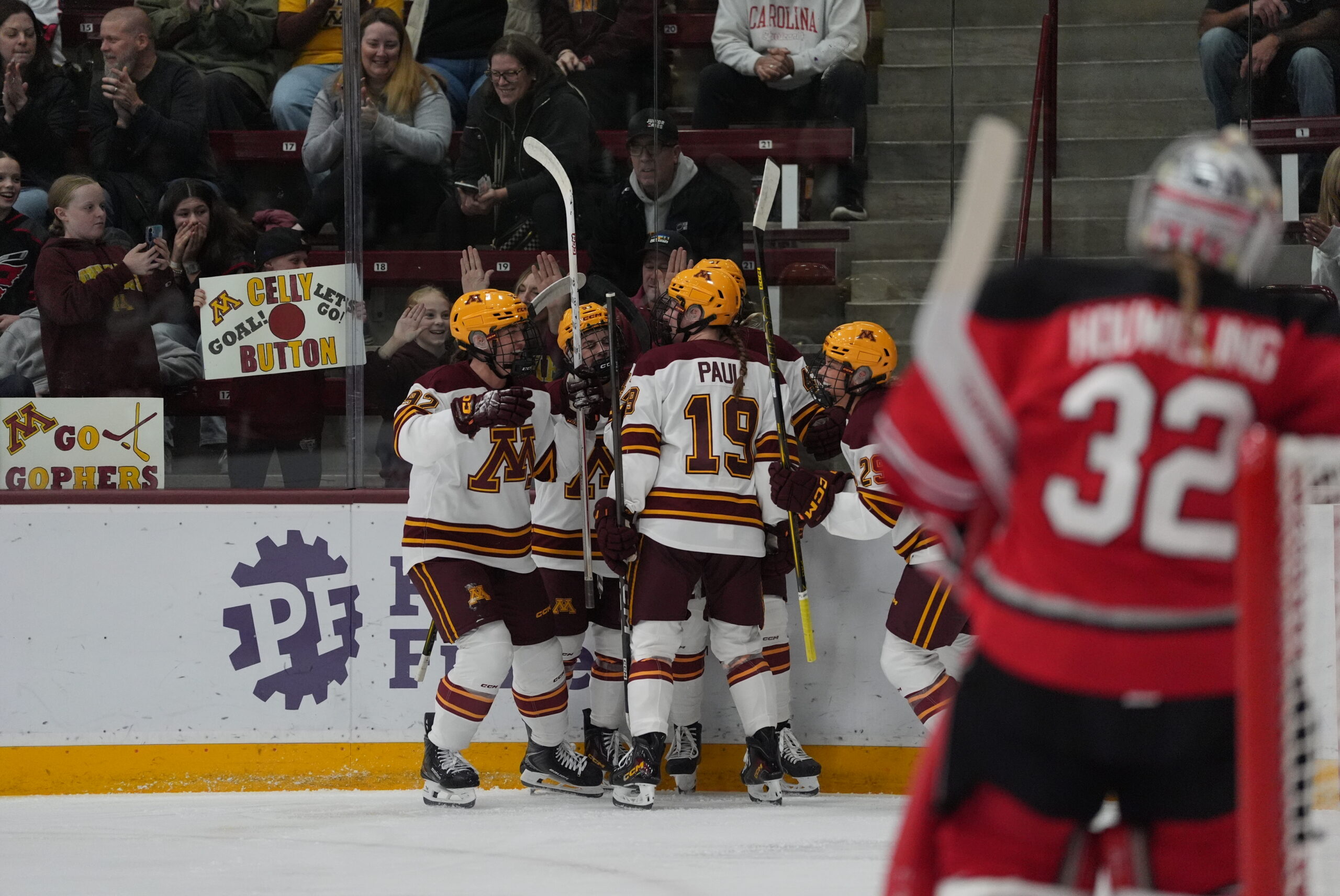 A hockey team in maroon and white jerseys celebrates on the ice, with fans cheering in the stands and a goalie in red visible in the foreground.