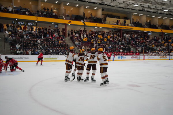 A group of ice hockey players in white and maroon uniforms celebrate on the ice during a game, with spectators watching from the stands.