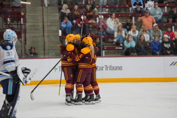 A group of ice hockey players in maroon and gold uniforms embrace on the ice after a play, with a goalie in white and blue gear nearby and spectators watching from the stands.