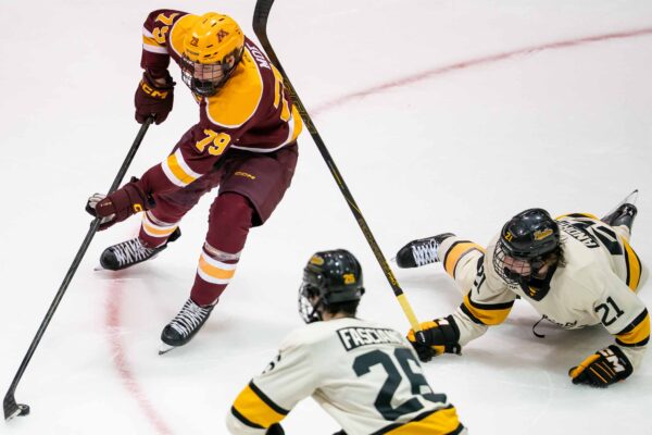 A hockey player in a maroon and gold uniform skates while two opponents in white and yellow gear attempt to defend him on the ice.